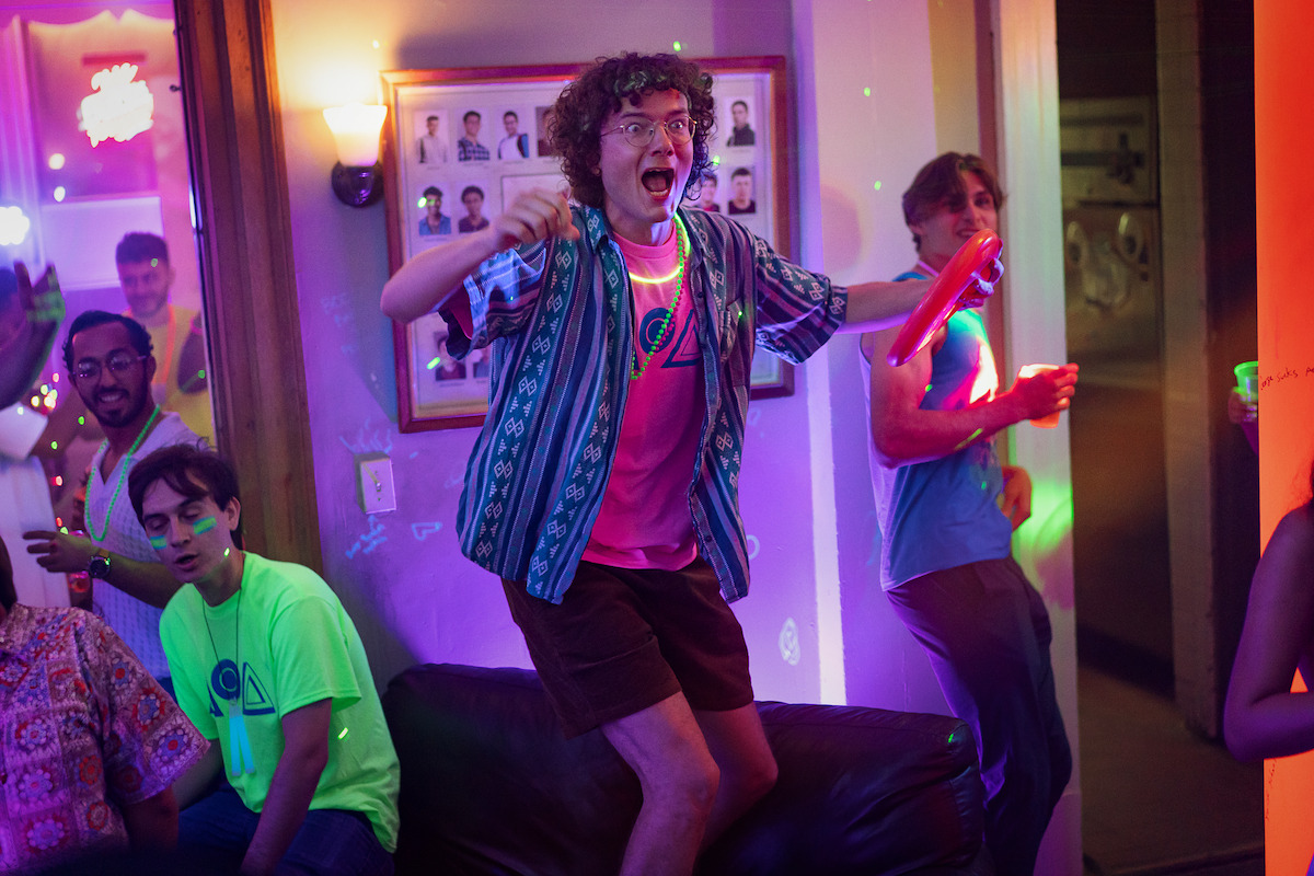 Group of young adults at a lively party with neon lights and glow sticks, one person excitedly standing on a couch, others sitting or dancing, colorful atmosphere and casual clothing in a home setting.