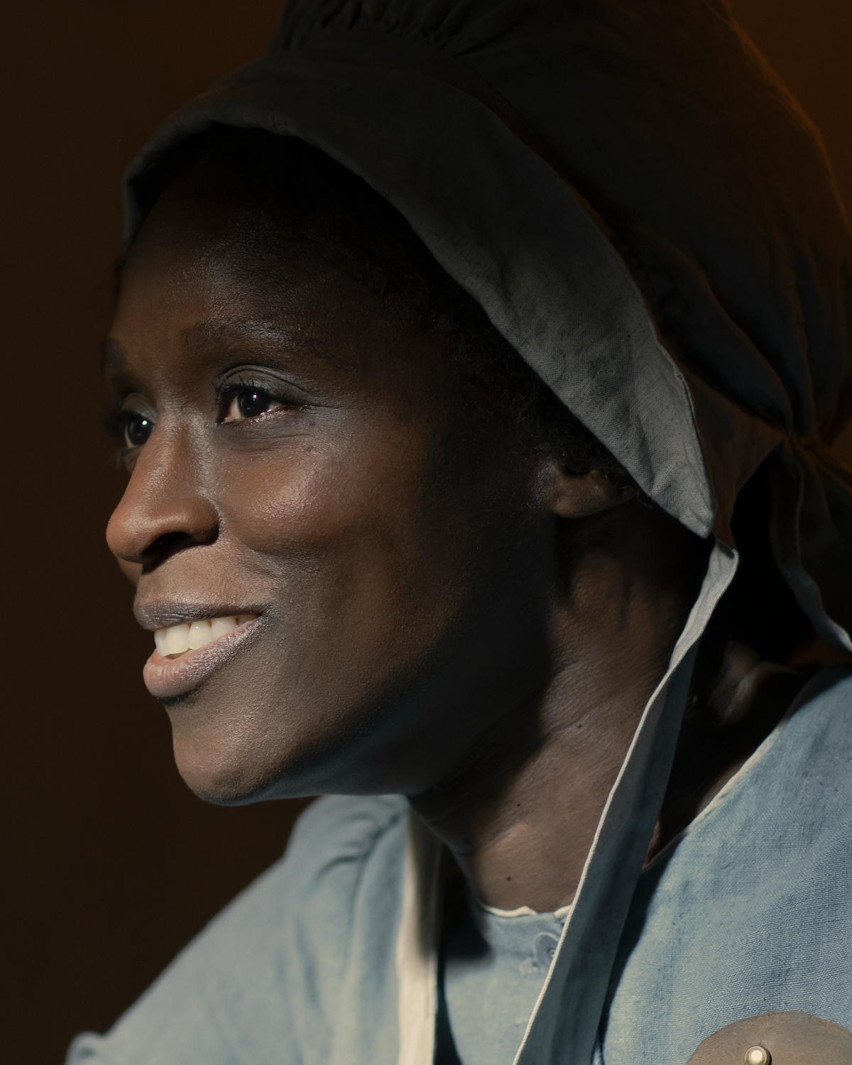 A woman wearing a historical bonnet and blue period clothing smiles warmly, shown in close-up lighting indoors, suggesting a historical or dramatic setting.