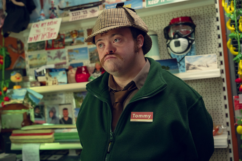 Man wearing a Sherlock Holmes-style hat and green jacket with a "Tommy" name tag standing behind the counter in a cluttered souvenir shop filled with postcards, snorkel masks, and various trinkets.