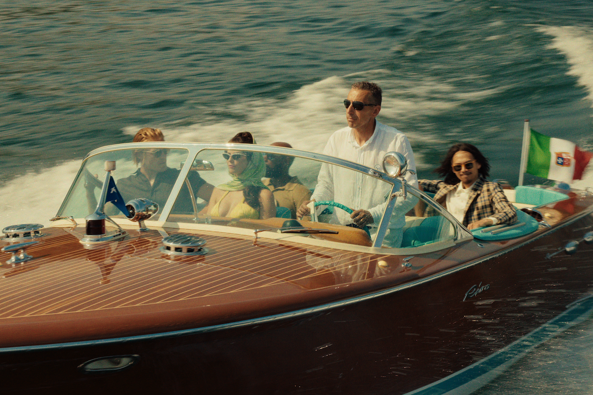 Four people ride in a classic wooden speedboat on open water, one man steering, others seated, with stylish attire and sunglasses, Italian and blue flags visible, creating a relaxed, luxurious atmosphere under sunny skies.