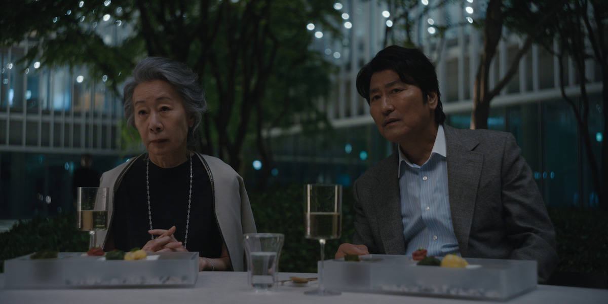 An elderly woman and a middle-aged man sit at an outdoor table in a modern, tree-filled courtyard at night, with glasses of wine, water, and plated food in front of them, surrounded by softly lit buildings.