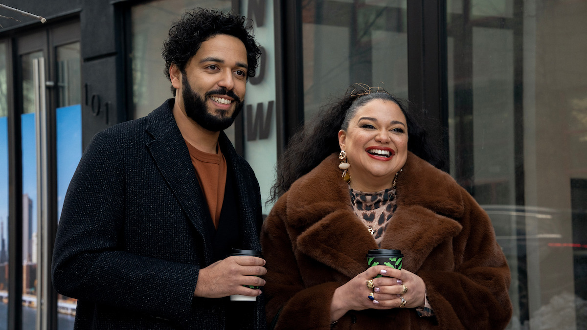 Two people smiling and holding coffee cups, standing outside a modern urban building. The man wears a dark coat and the woman is in a brown fur coat with bold accessories. City reflections are visible in the glass behind them.