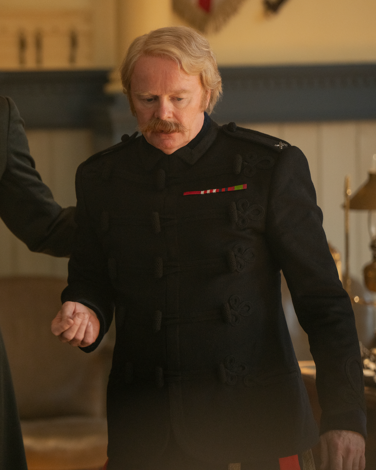 Man with blonde hair and mustache wearing a formal dark military-style uniform with medals, standing indoors in a warmly lit room with wood paneling and vintage furniture in the background.