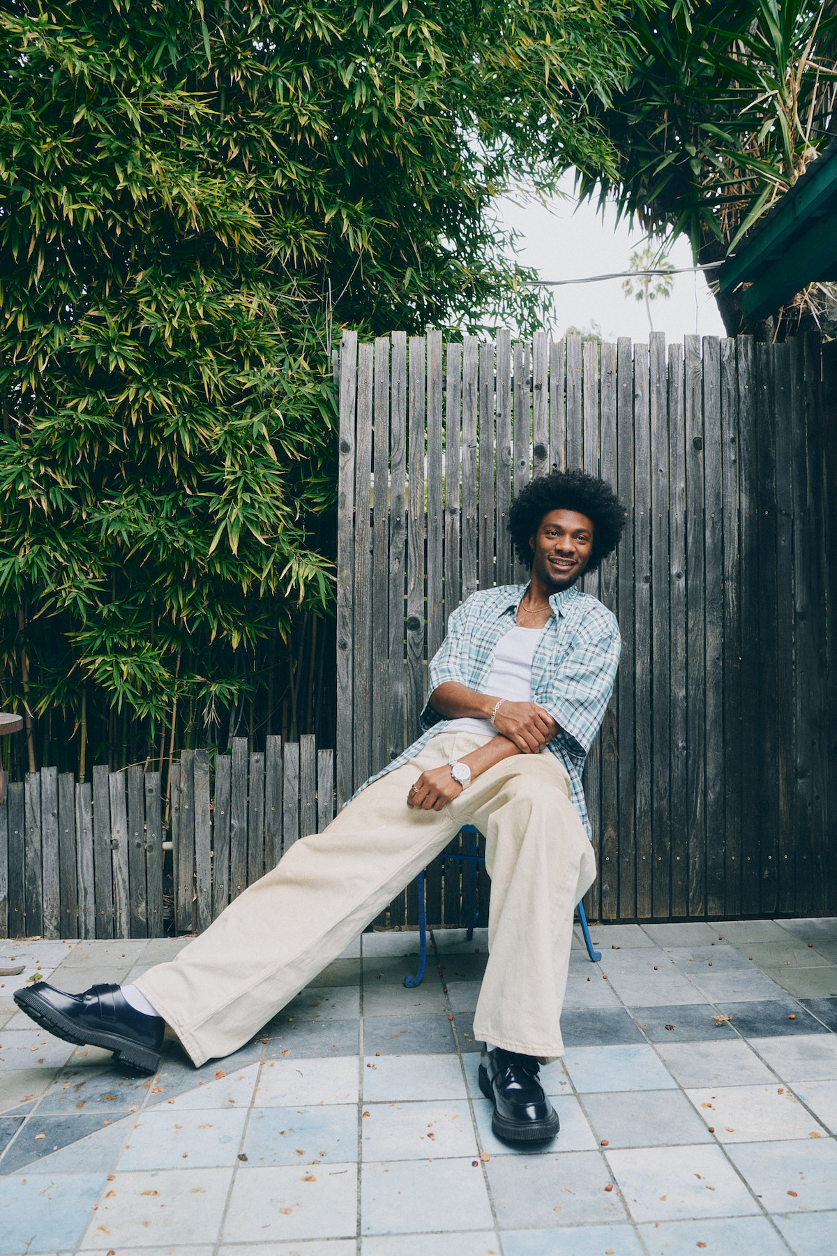 Man with curly hair sitting casually on a blue chair outside on a tiled patio, wearing a plaid shirt, white t-shirt, and beige pants, with a wooden fence and lush green bamboo in the background. Relaxed outdoor atmosphere.