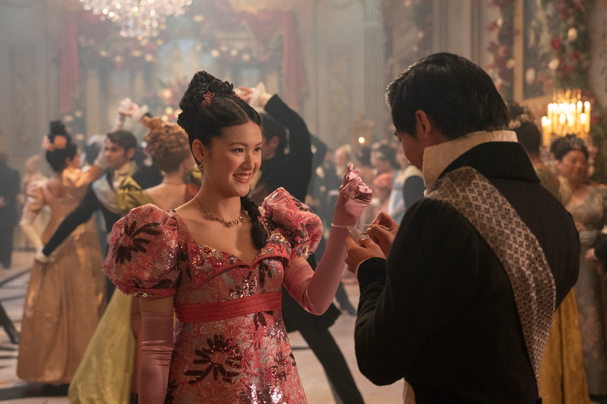 A woman in an elaborate pink gown dances with a man in formal period clothing at a grand ballroom event, surrounded by other elegantly dressed guests and ornate decorations.