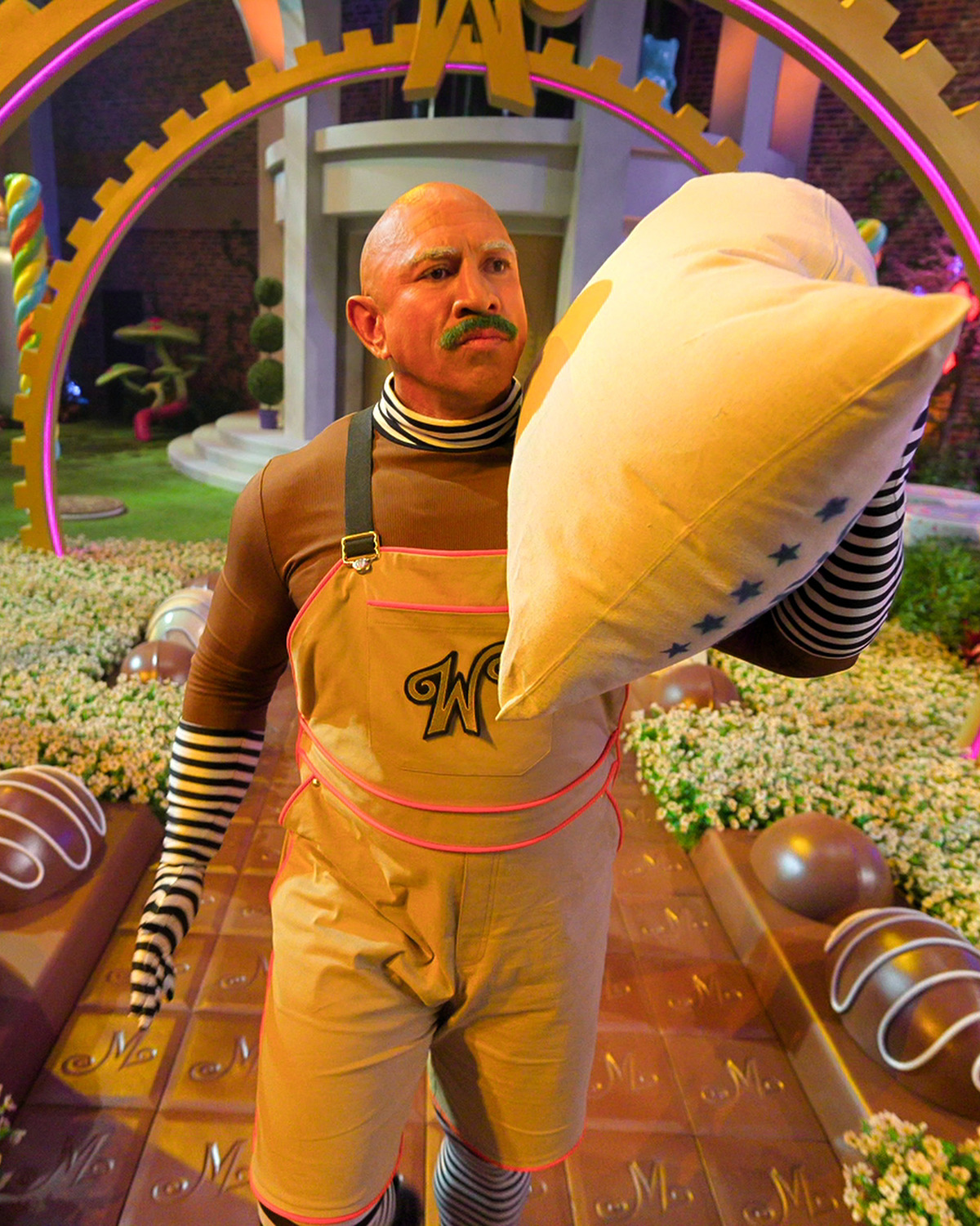 A person in a whimsical costume with striped sleeves and a "W" logo holds a large pillow in a colorful, candy-themed garden with oversized chocolate and candy decorations, vibrant lighting, and a playful atmosphere.