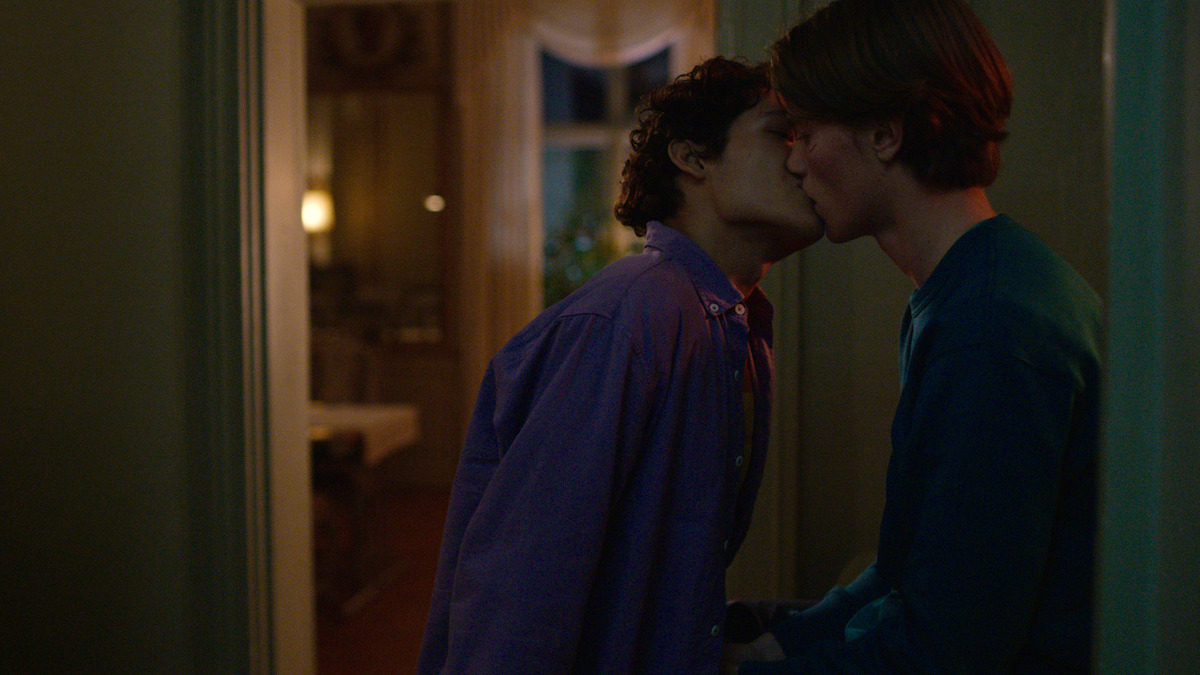 Two young people share a kiss in the doorway of a dimly lit room with warm lighting and a window in the background, creating an intimate and cozy atmosphere.