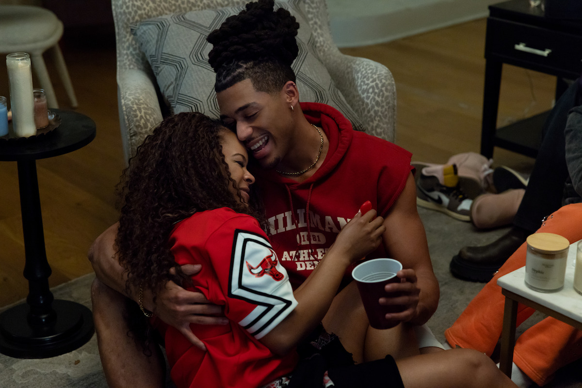 Two young people sit on a living room floor, laughing and hugging at a casual indoor gathering. Drinks, candles, and casual seating create a relaxed, cozy atmosphere.