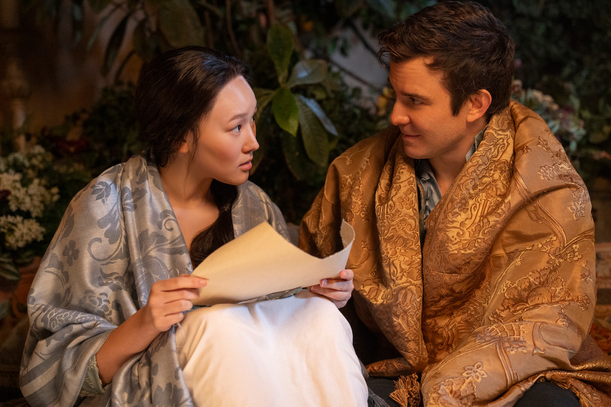 Two people sit closely together wrapped in ornate blankets, one holding a piece of paper. They are indoors with plants and soft lighting, engaged in a serious conversation.