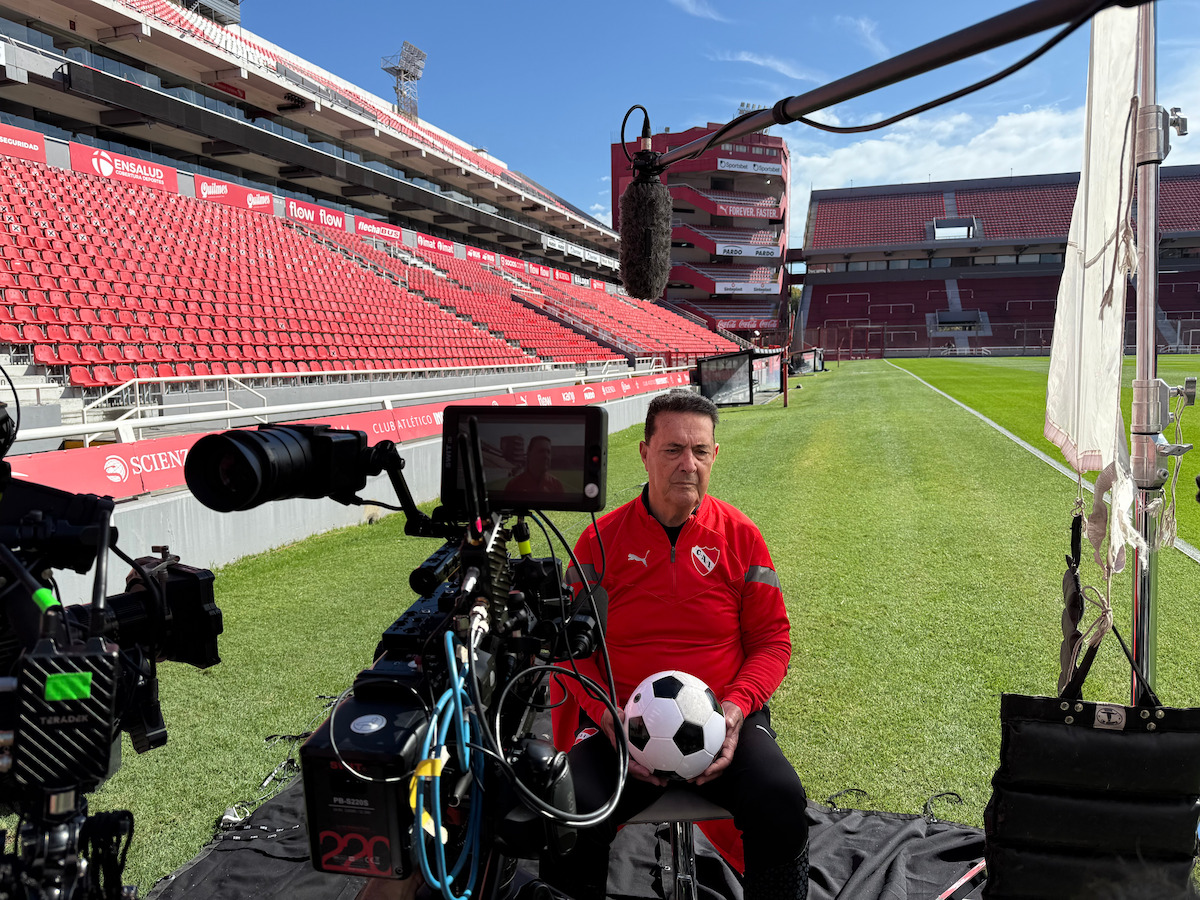 Man in red sports jacket holding soccer ball, sitting on chair for interview on stadium sideline, surrounded by professional video cameras, microphone, and lighting equipment, with empty red stadium seats and soccer field in background.