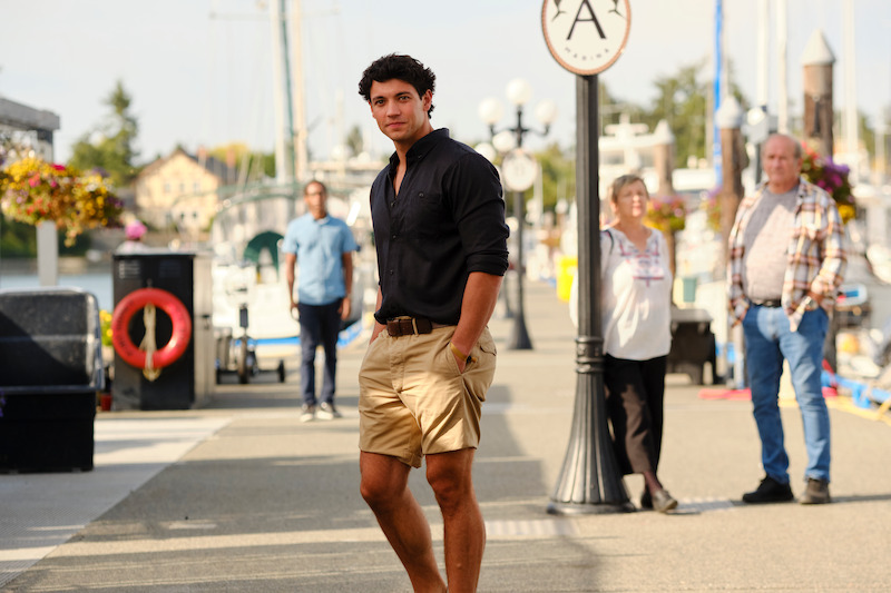 Young man in black shirt and khaki shorts walking on a marina dock, with boats, flowers, and people in the background on a sunny day.
