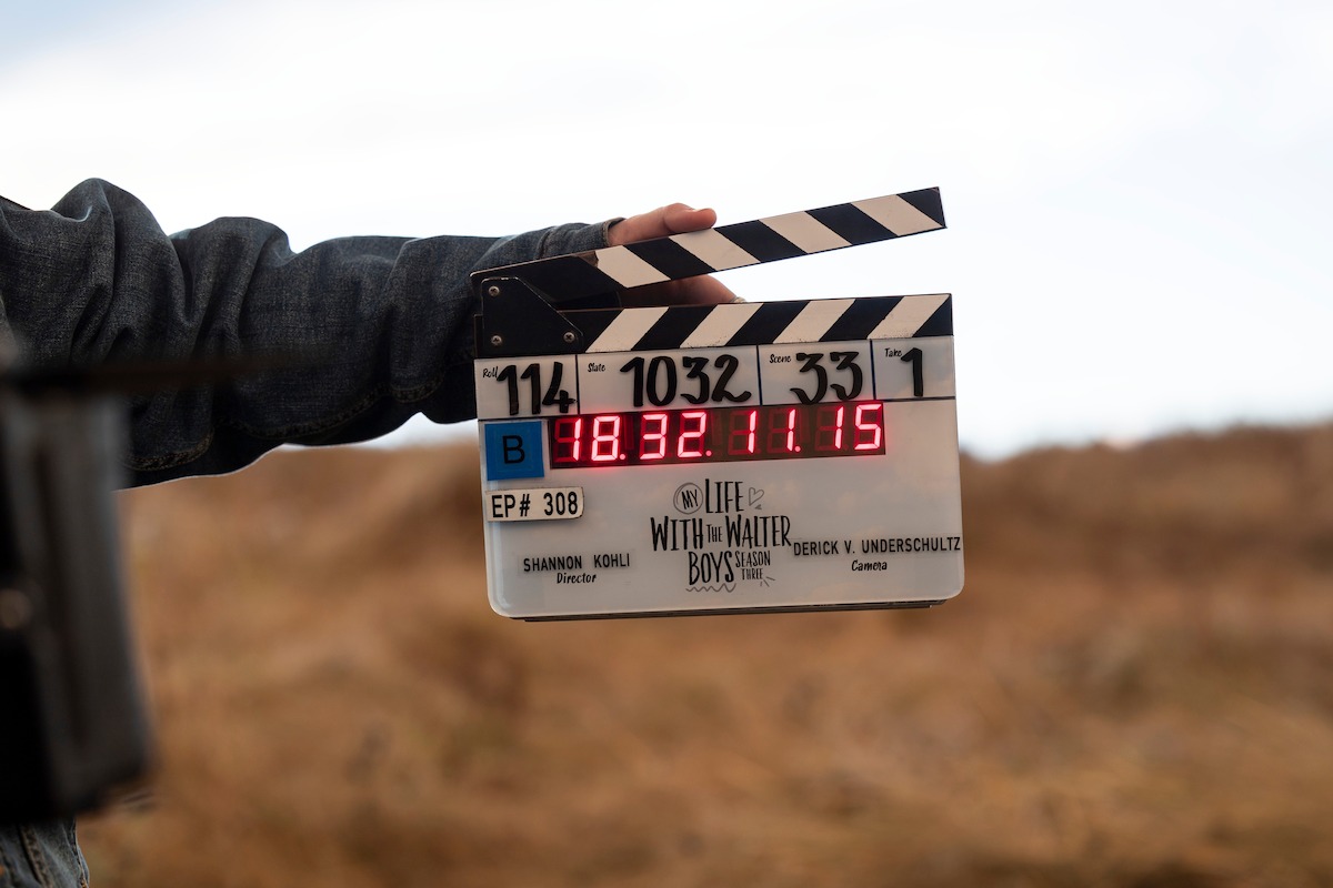A person holding a film clapperboard with digital time display outdoors in a field, indicating a movie or TV production scene being shot in a natural, grassy setting.