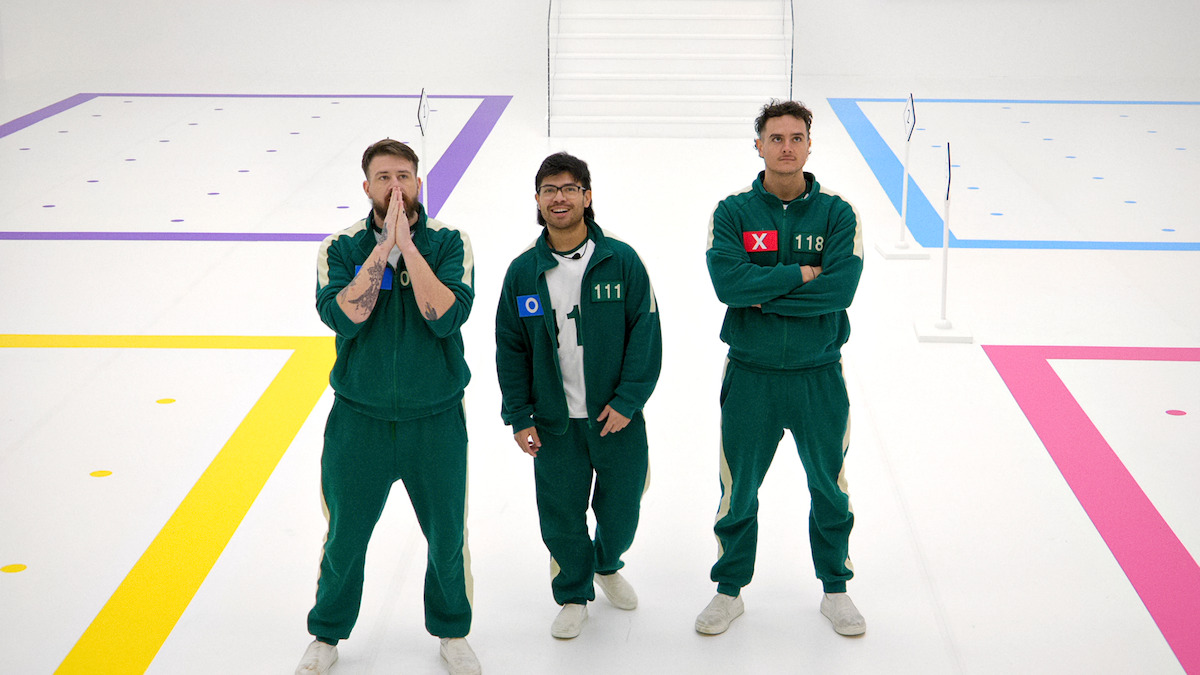 Three men in green tracksuits stand on a brightly lit, colorful game set with geometric markings. The mood is tense and competitive, suggesting a serious moment in a challenge or competition.