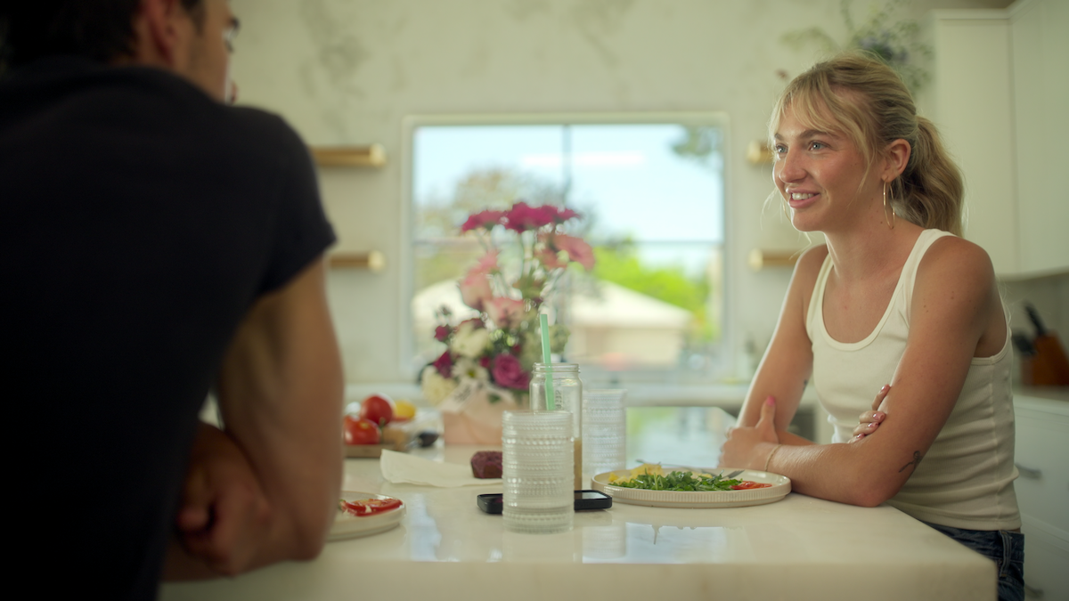 Two people sit at a kitchen counter with plates of food, glasses, and flowers on display. They are talking in a bright, modern kitchen with sunlight coming through a window.