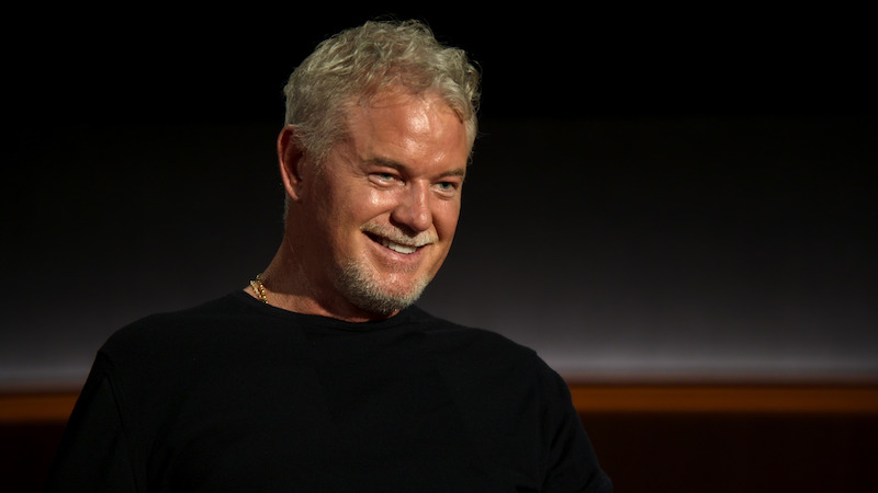Eric Dane sits against a black backdrop wearing a black shirt.