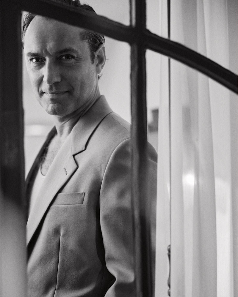 Man in a suit stands indoors by a window with sheer curtains, looking at the camera, captured in black and white with soft natural lighting.