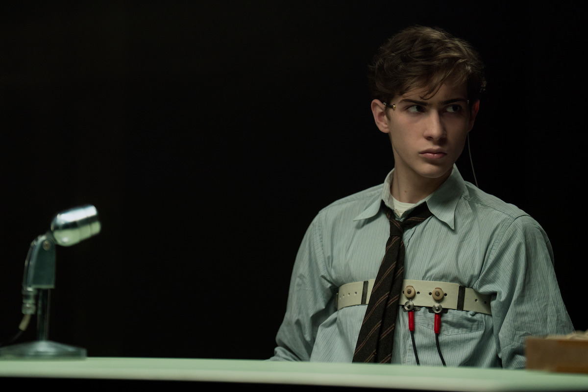 Young man in formal clothes sits at a desk with a polygraph machine attached, microphone nearby, dark interrogation room setting, tense expression.