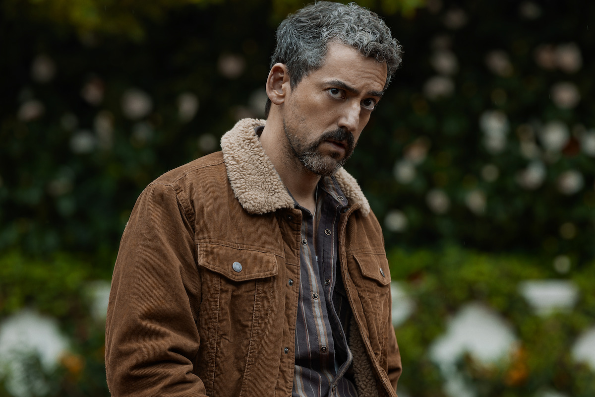 Man with gray hair and beard wearing a brown jacket with a shearling collar, standing outdoors in a garden or park setting with greenery and blurred flowers in the background, looking pensively to the side.