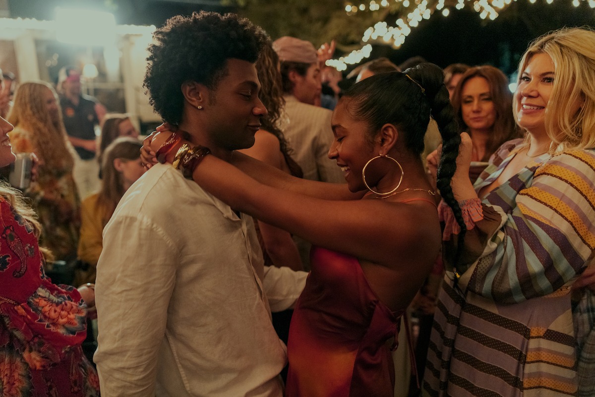 A couple dances closely at an outdoor party surrounded by smiling guests in colorful outfits under string lights at night.