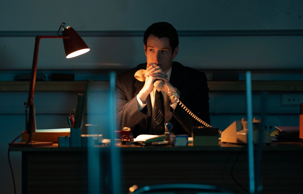 A man in a suit sits alone at a desk in a dimly lit office, holding a telephone receiver, with a red desk lamp providing focused light on the work surface.