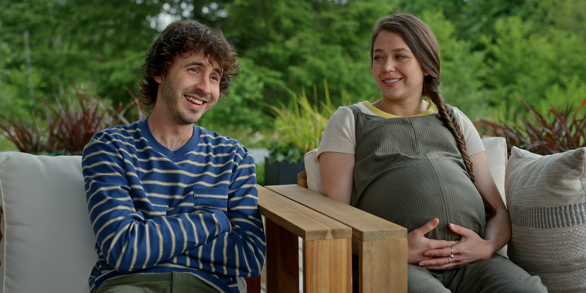 A man and a pregnant woman sit on a patio sofa outdoors, surrounded by greenery, smiling and talking together in a relaxed garden setting.