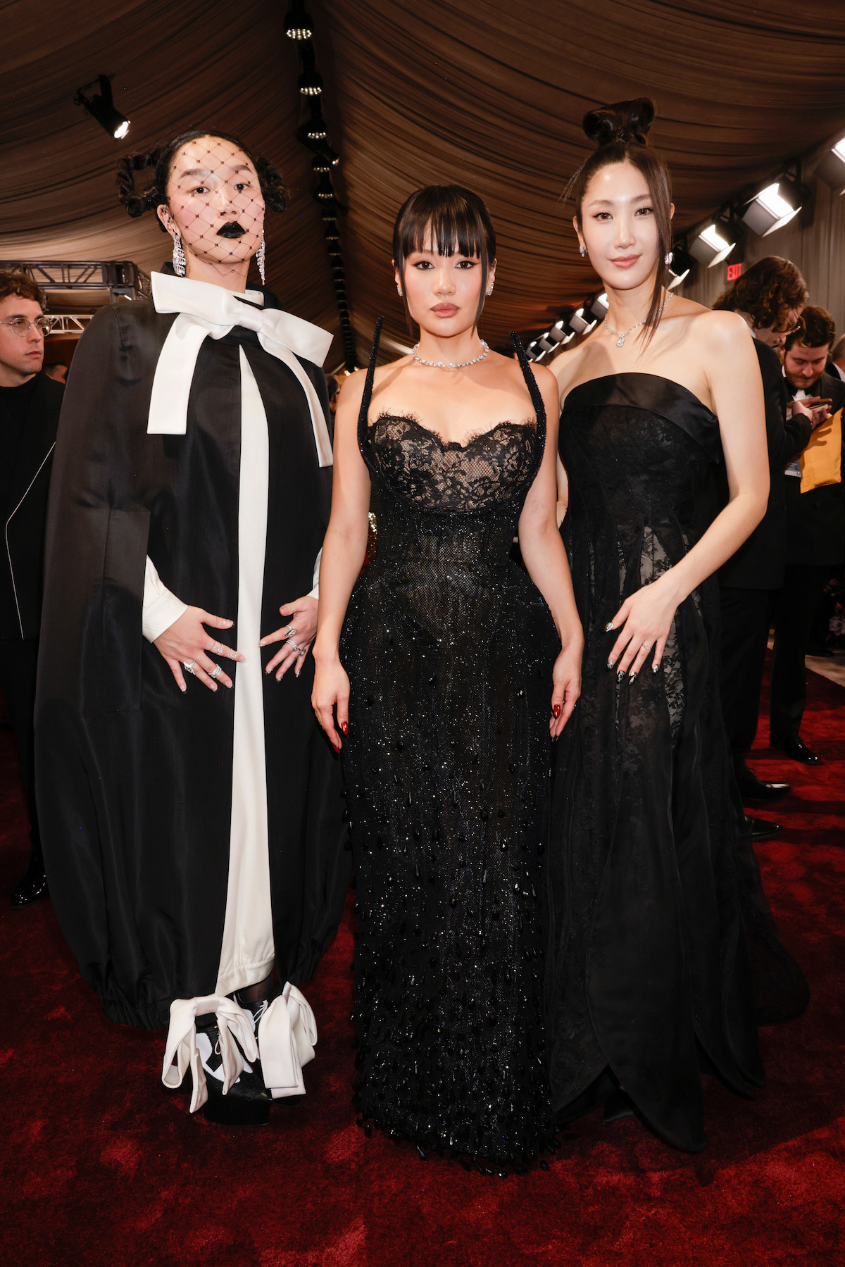 Three people in formal black attire pose on a red carpet under a draped ceiling with lights; one in an avant-garde ensemble with face netting, the others in elegant black dresses, amid event attendees.