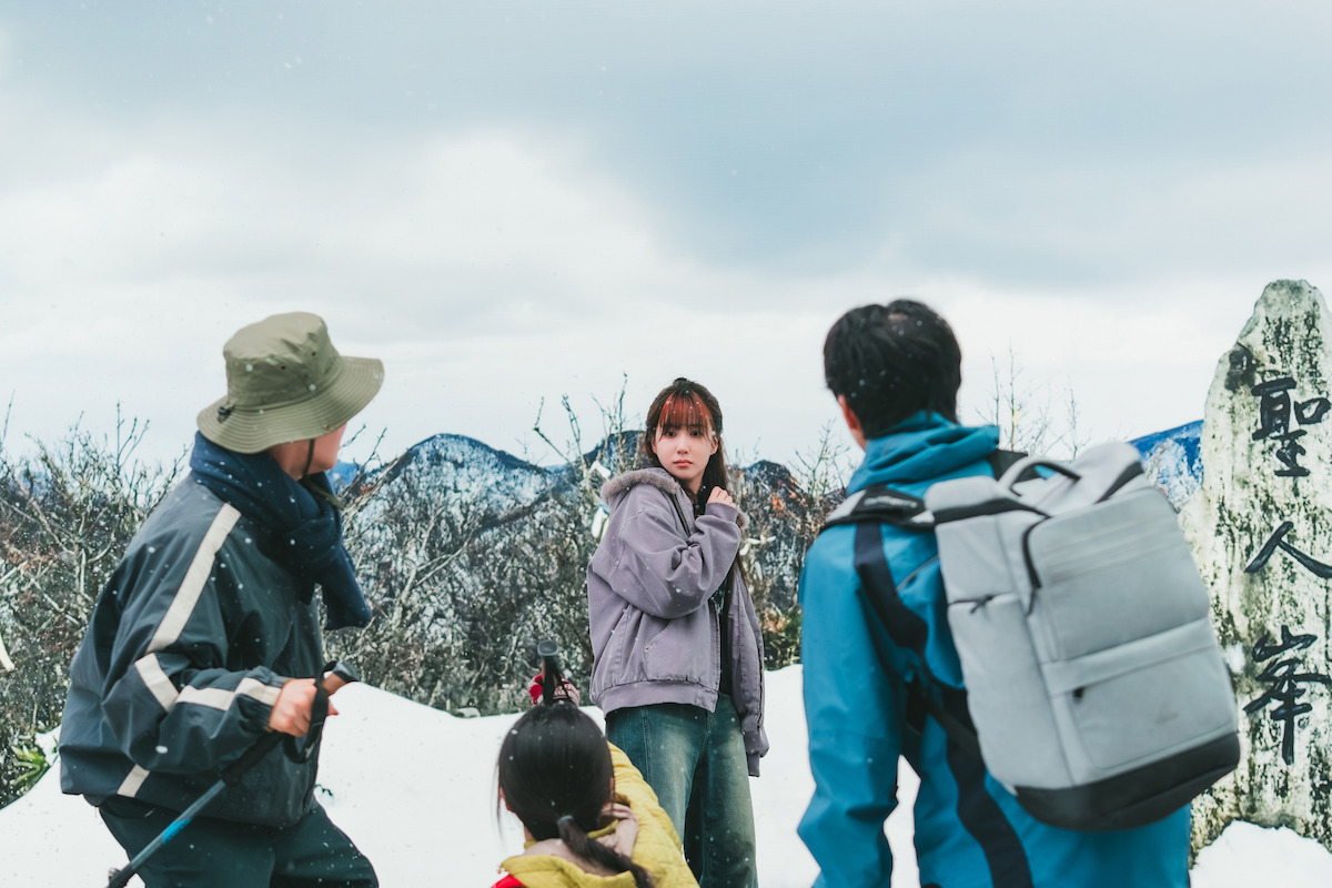 Four people, including a woman in a purple jacket, stand on a snowy mountain with hiking gear. They are outdoors near a stone marker with Japanese writing, surrounded by winter trees and cloudy skies.