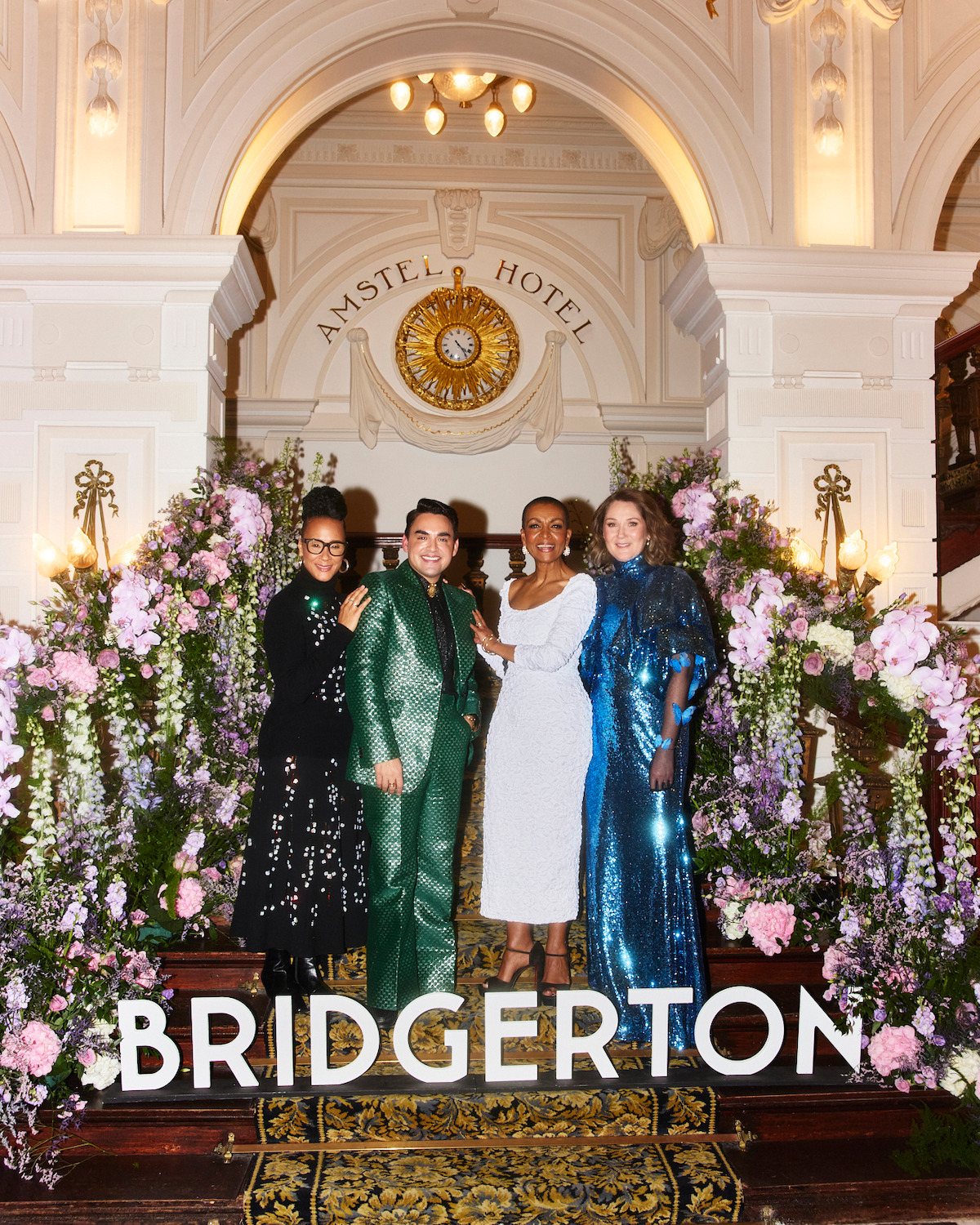 Four people dressed elegantly pose on a grand staircase decorated with flowers in an ornate hall, with "Bridgerton" text at the bottom, evoking a formal, historical setting.