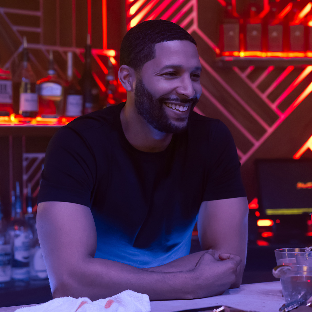 Norman Nixon Jr. as Prof. Isaiah Baptiste leans on a bar counter in a trendy, neon-lit bar with bottles and glasses in the background, creating a lively nightlife atmosphere.
