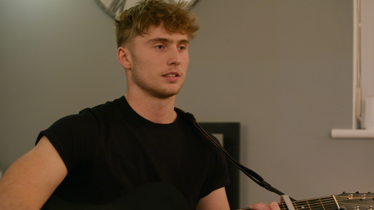 Young man in a casual black shirt playing acoustic guitar indoors with neutral walls; calm, focused mood in a cozy home environment.