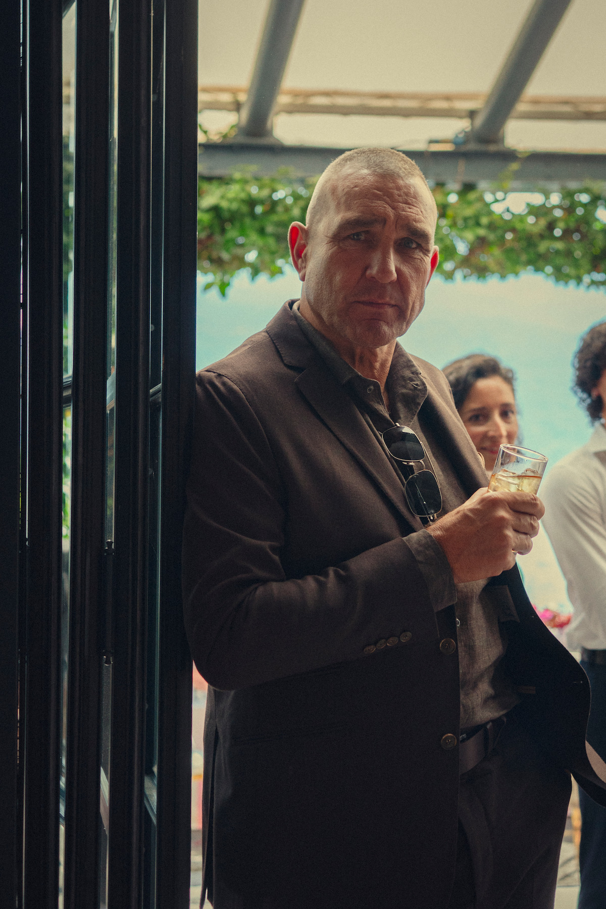 Man in a brown suit holding a drink, standing indoors near a window with greenery outside, with people socializing in the background during daytime.