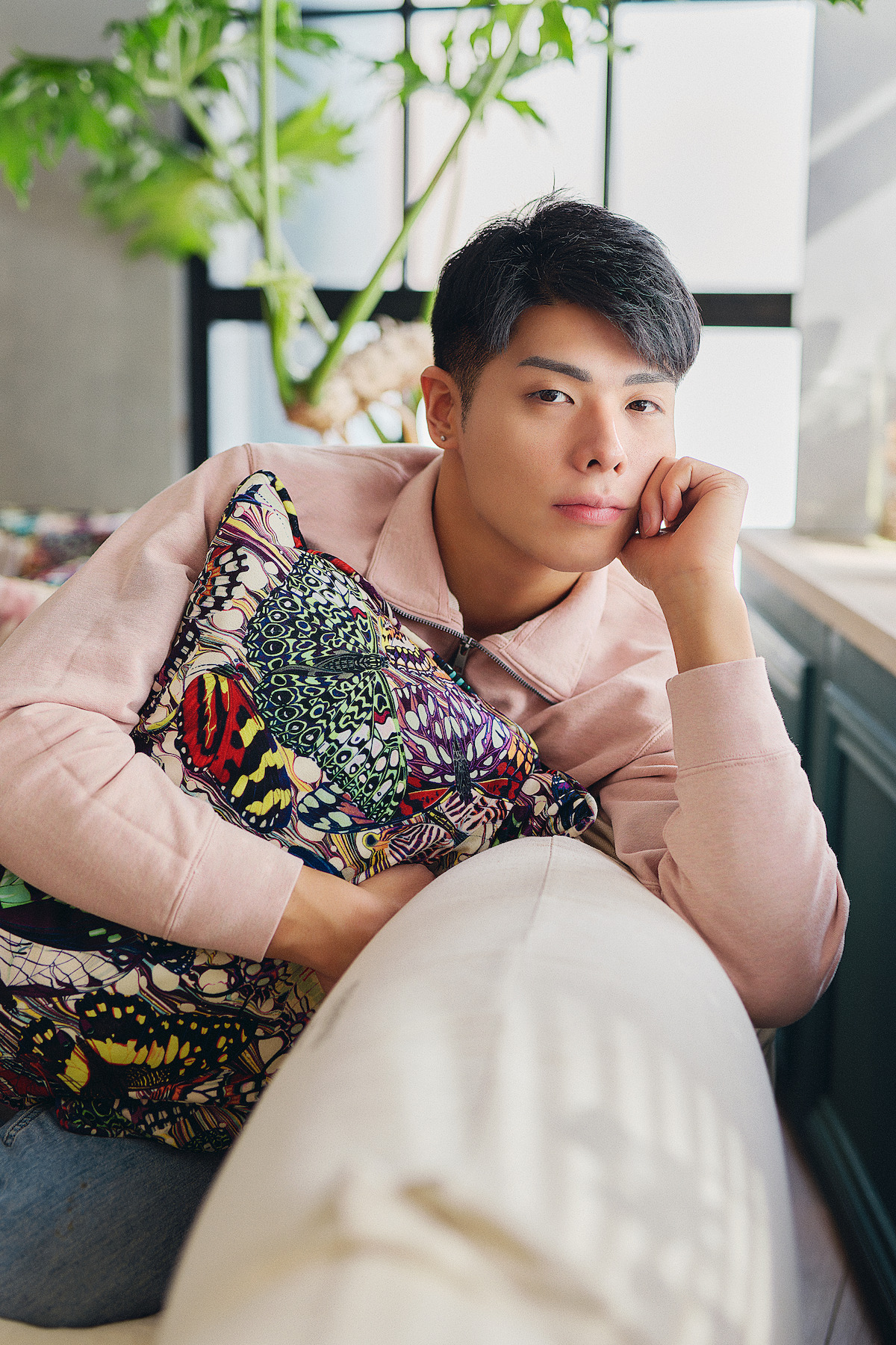 A young man in a pink sweatshirt sits on a sofa, resting his head on one hand and holding a colorful cushion in a bright, modern room with large windows and green plants, creating a calm and relaxed mood.