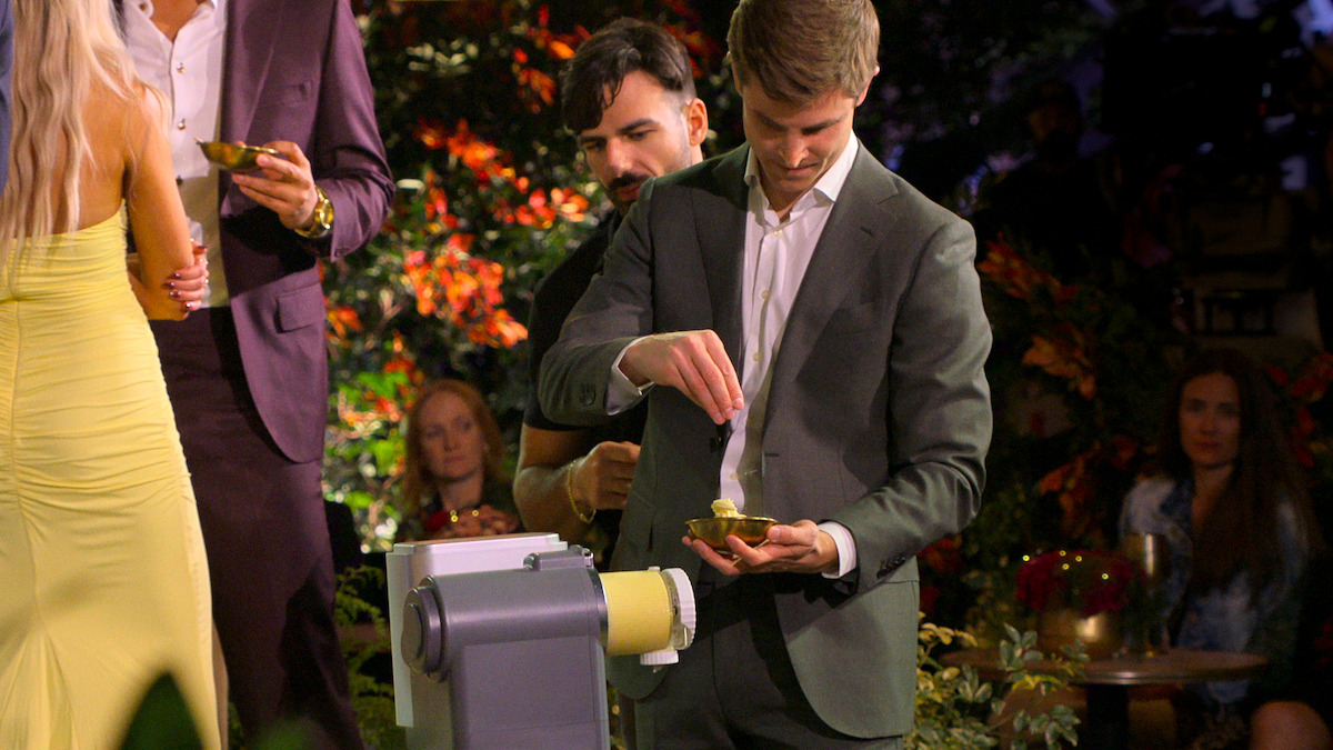 Man in suit adding topping to plate at a buffet in a festive outdoor garden setting, people socializing around him, warm lighting creates a cheerful and elegant atmosphere.