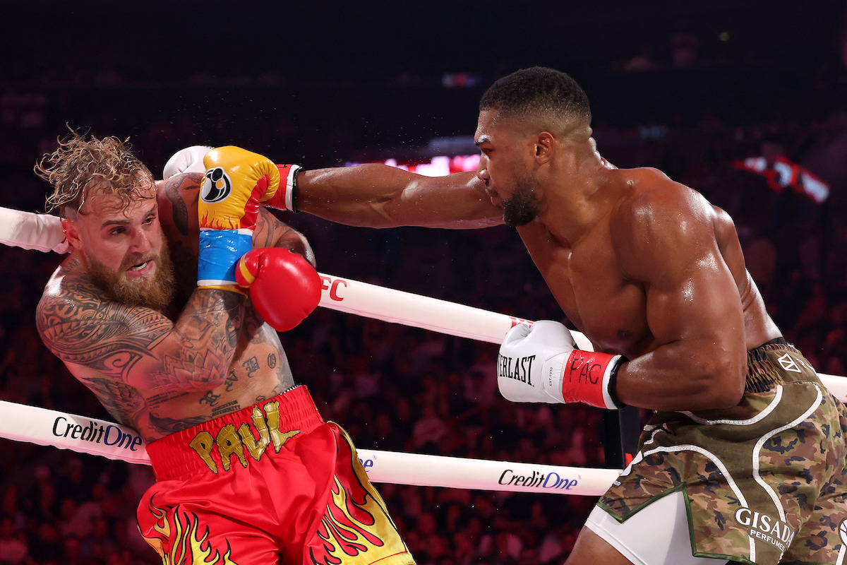 Anthony Joshua fighting Jake Paul in a brightly lit boxing ring, wearing gloves and sports gear, with bright arena lights and a crowd in the background.
