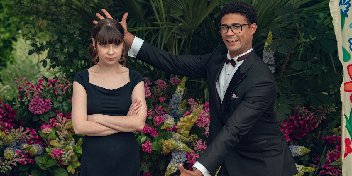 A man in a tuxedo playfully poses next to a woman in a black dress who stands with arms crossed and a serious expression, in front of colorful flowers and greenery outdoors.
