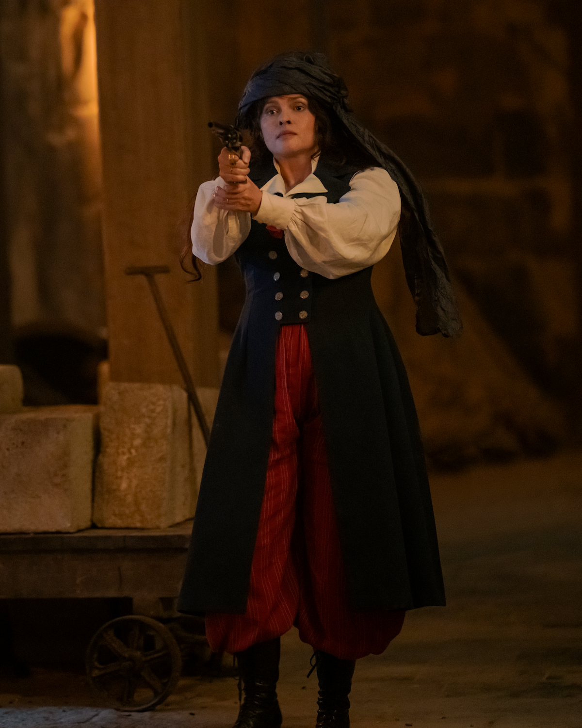 Person in historical costume with a headscarf and long vest, aiming a pistol indoors, standing on a stone floor near large blocks and a wooden cart, in dim, warm lighting suggestive of a historical drama or reenactment setting.