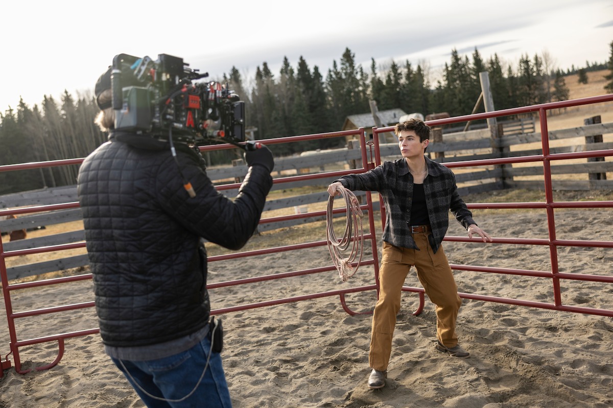 A camera operator films a young person holding a lasso in a fenced outdoor corral, surrounded by trees and rural landscape under a cloudy sky.