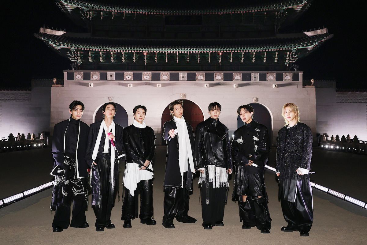 V, JIN, SUGA, RM, JUNG KOOK, J-HOPE, and JIMIN dressed in fashionable black and white outfits stand outdoors at night in front of an ornate, traditional East Asian building with arches and a large decorative roof.