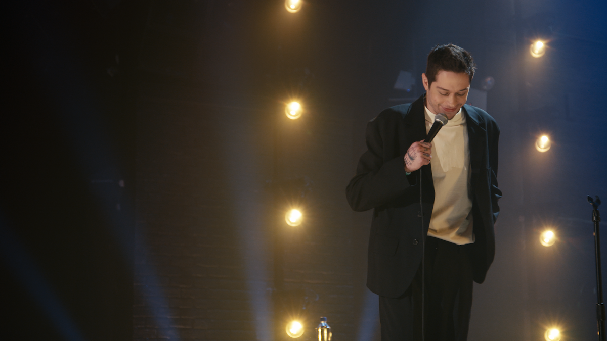Comediante en el escenario con un traje oscuro sosteniendo un micrófono, actuando bajo cálidas luces del escenario colocadas contra una pared de ladrillos, creando una atmósfera de comedia de stand-up íntima y centrada.