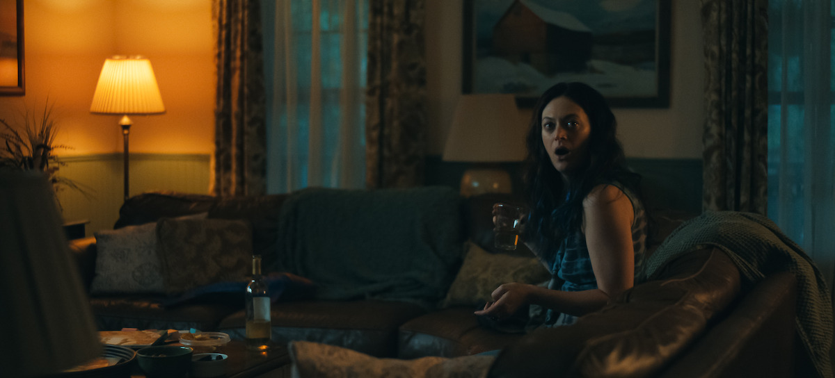 A woman sits on a couch in a dimly lit cozy living room at night, holding a glass of wine with her mouth open in surprise.