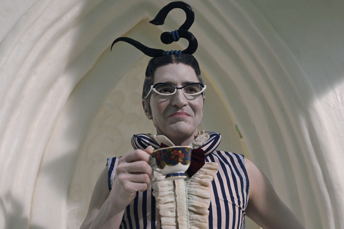 Person with elaborate black hairstyle shaped like the number three, wearing unique glasses and a striped costume, holds a teacup, standing in front of a light, curved architectural background.