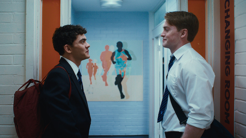 Two students in uniforms stand facing each other in a brightly lit school hallway outside a changing room, with a colorful mural of runners on the back wall.