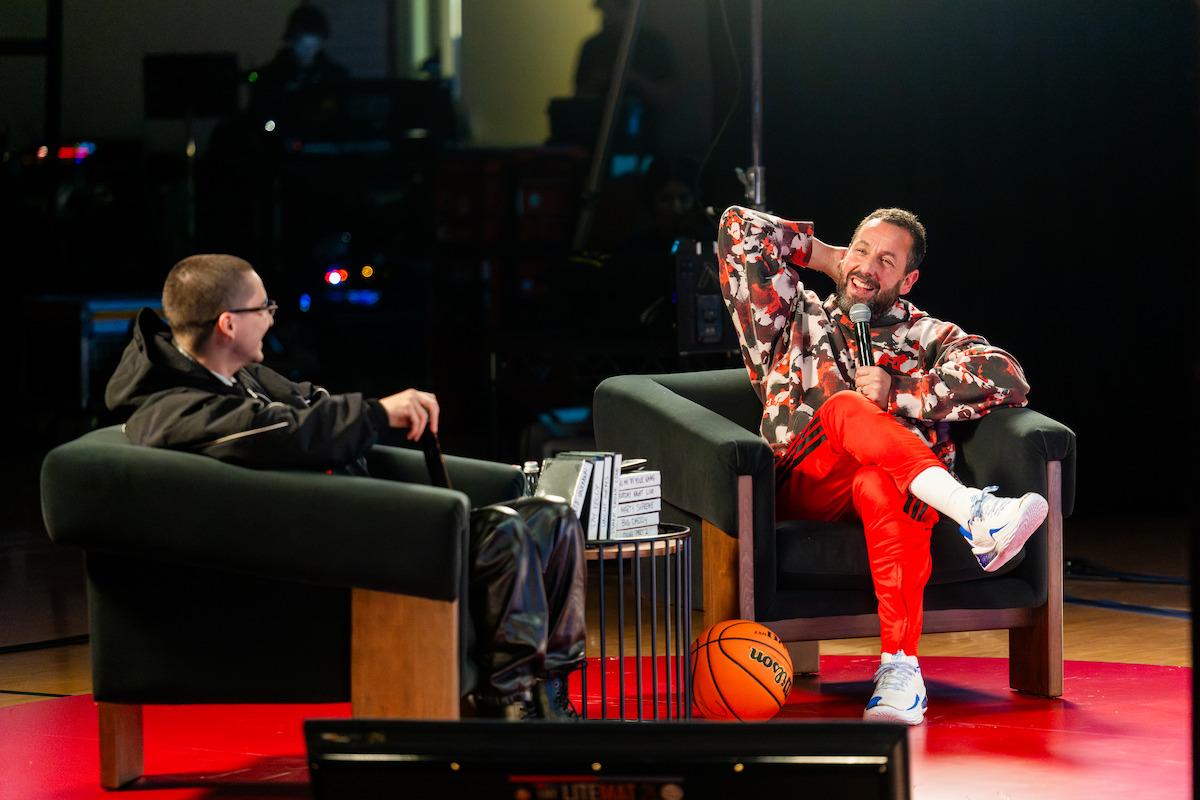 Two men sit in armchairs on a stage having a lively conversation. One is relaxed and smiling, wearing bright red pants, with a basketball at their feet. The environment is casual with a warm, friendly mood.