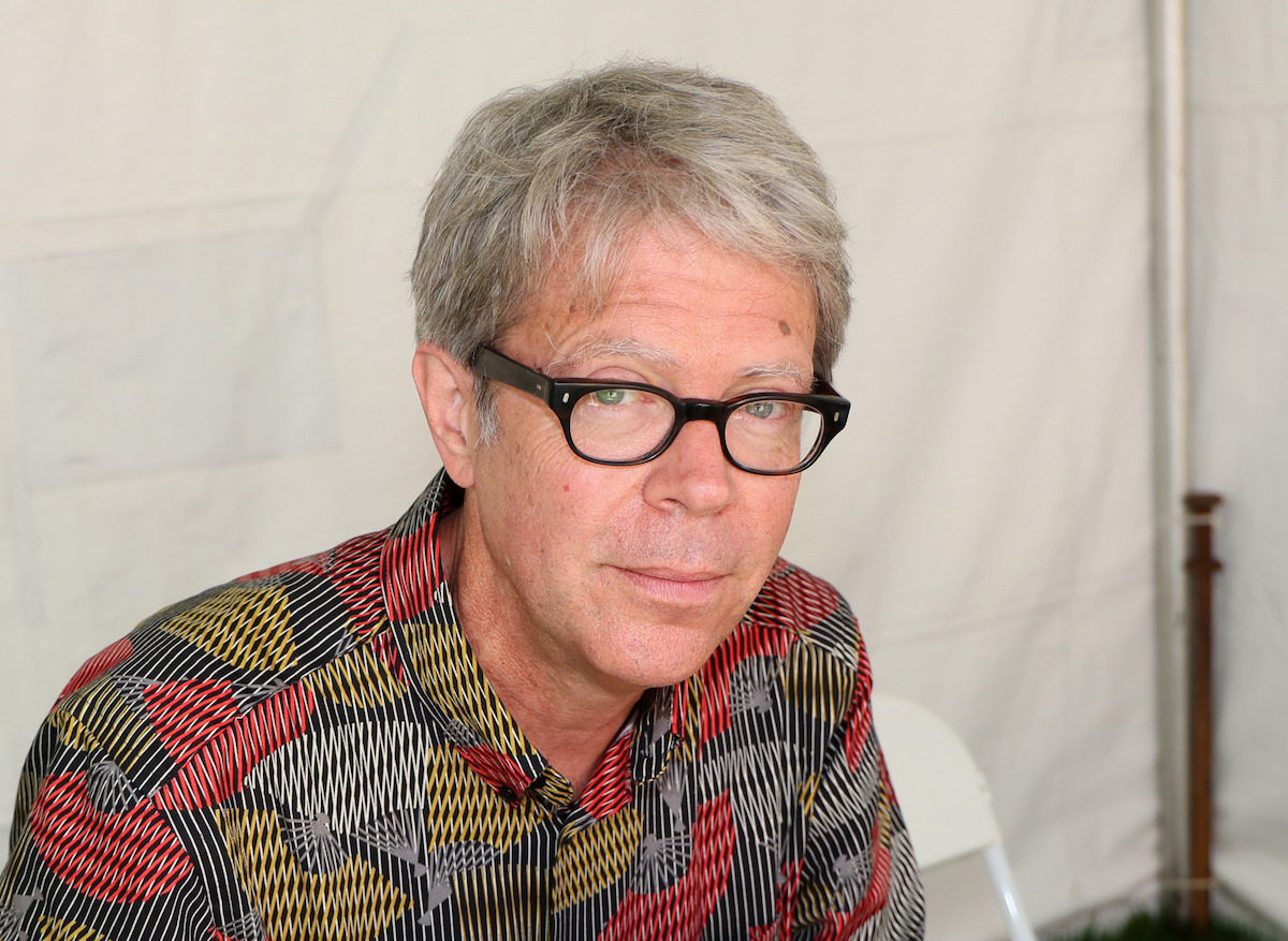 Middle-aged man with gray hair and glasses wearing a colorful patterned shirt, sitting indoors against a white tent backdrop, looking at the camera.