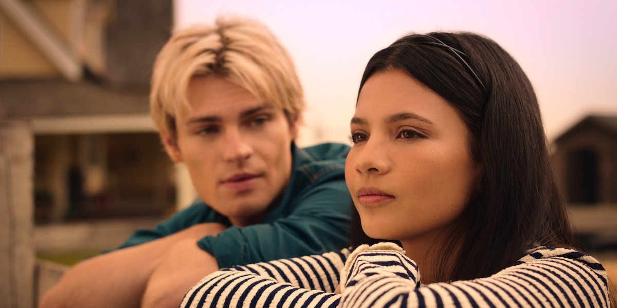 Two young adults sitting close outdoors, with a house in the background. The woman in the foreground looks thoughtful, wearing a striped sweater, while the man behind her watches her with a gentle expression in soft, warm lighting.
