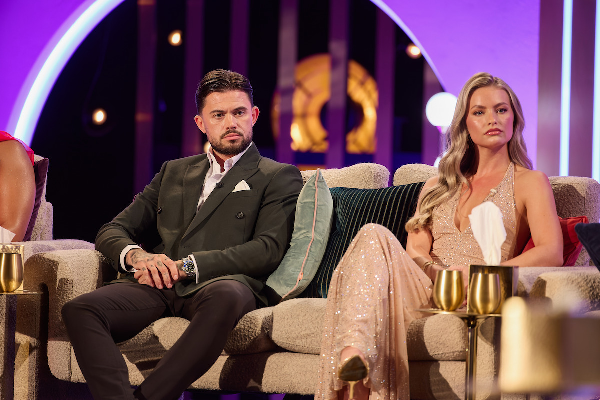 Man in a dark suit and woman in a sparkly gown sitting on a modern sofa at a formal indoor event with stylish lighting, gold decor, and elegant tableware, both appearing serious or focused.