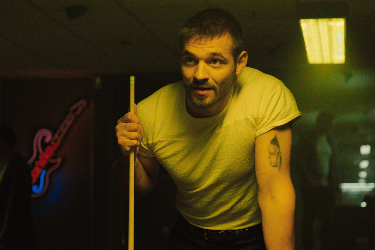 Man in white t-shirt holding pool cue, leaning over a pool table in a dimly lit room with neon guitar sign and overhead lights, focused and ready to play.