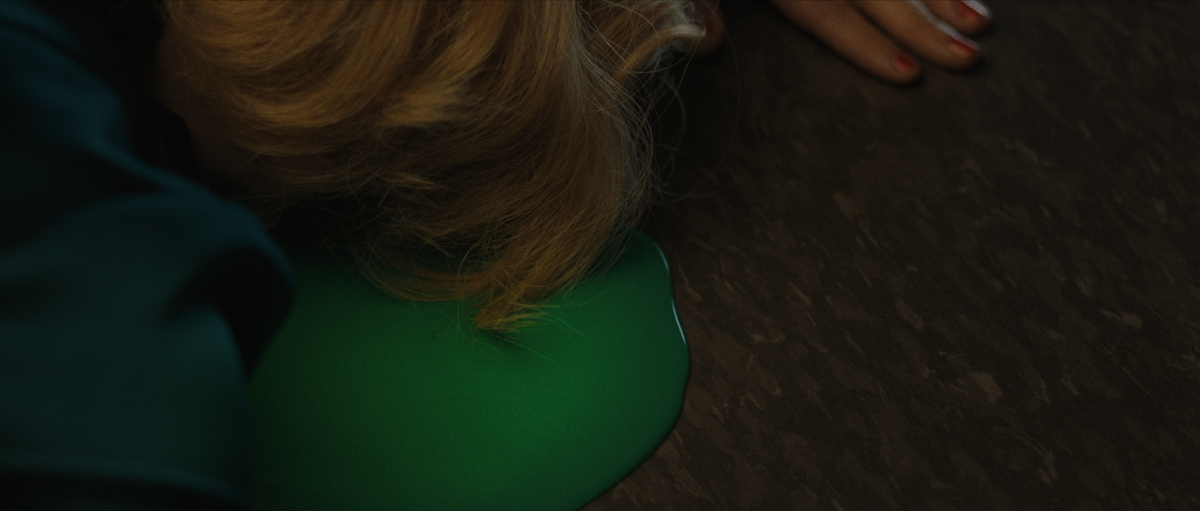 A person with blonde hair is lying face-down on a dark floor, partially on a puddle of bright green liquid, with one hand visible and outstretched. The setting appears to be indoors with dim lighting.
