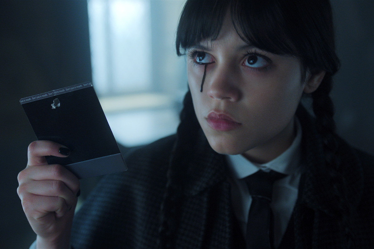 Jenna Ortega as Wednesday Addams holds a poloroid photograph and sheds a single black tear in ‘Wednesday’ Season 2.