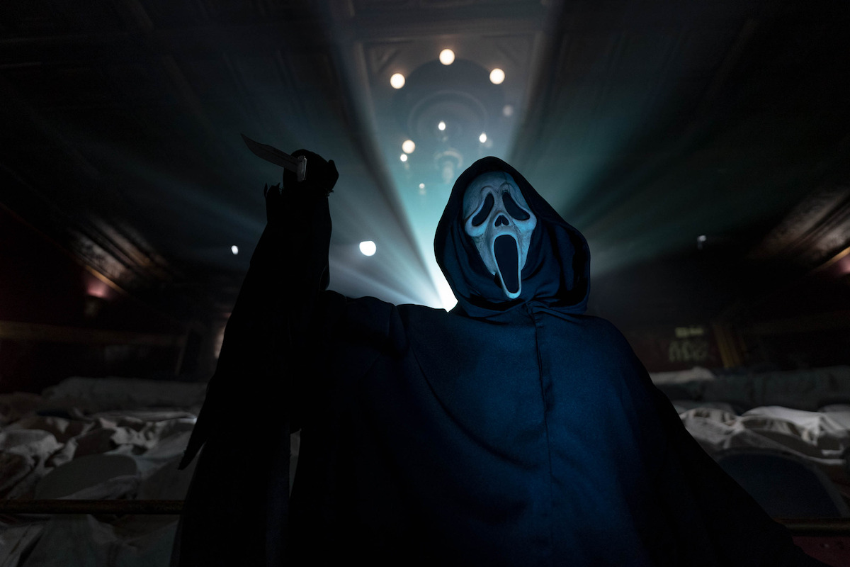 Person in a black cloak and ghostly white mask holding a knife, standing in a dramatic, dimly lit theater with rows of seats and light beams illuminating the background.